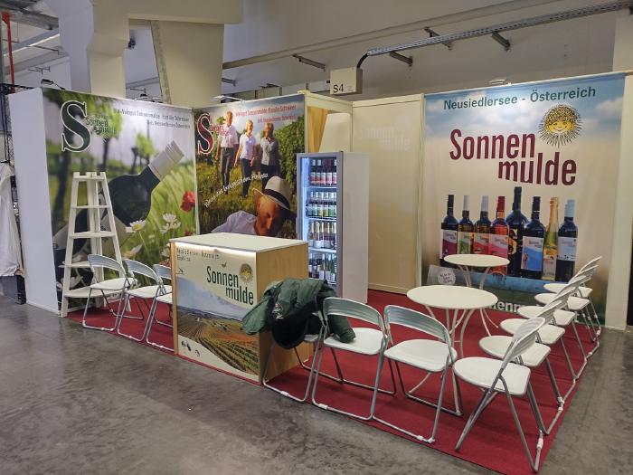 An exhibition stand with carpet, tables and chairs. At the aisle a desk with a printed picture and logo. In the background, walls with pictures of wine bottles and of people.