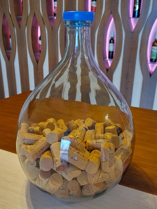 On a table is a glass balloon, a balloon-shaped glass bottle about 20 liters in size. It is filled to about half of the bellied part with a variety of wine corks. In the background there is a wall with niches in which wine bottles are displayed.